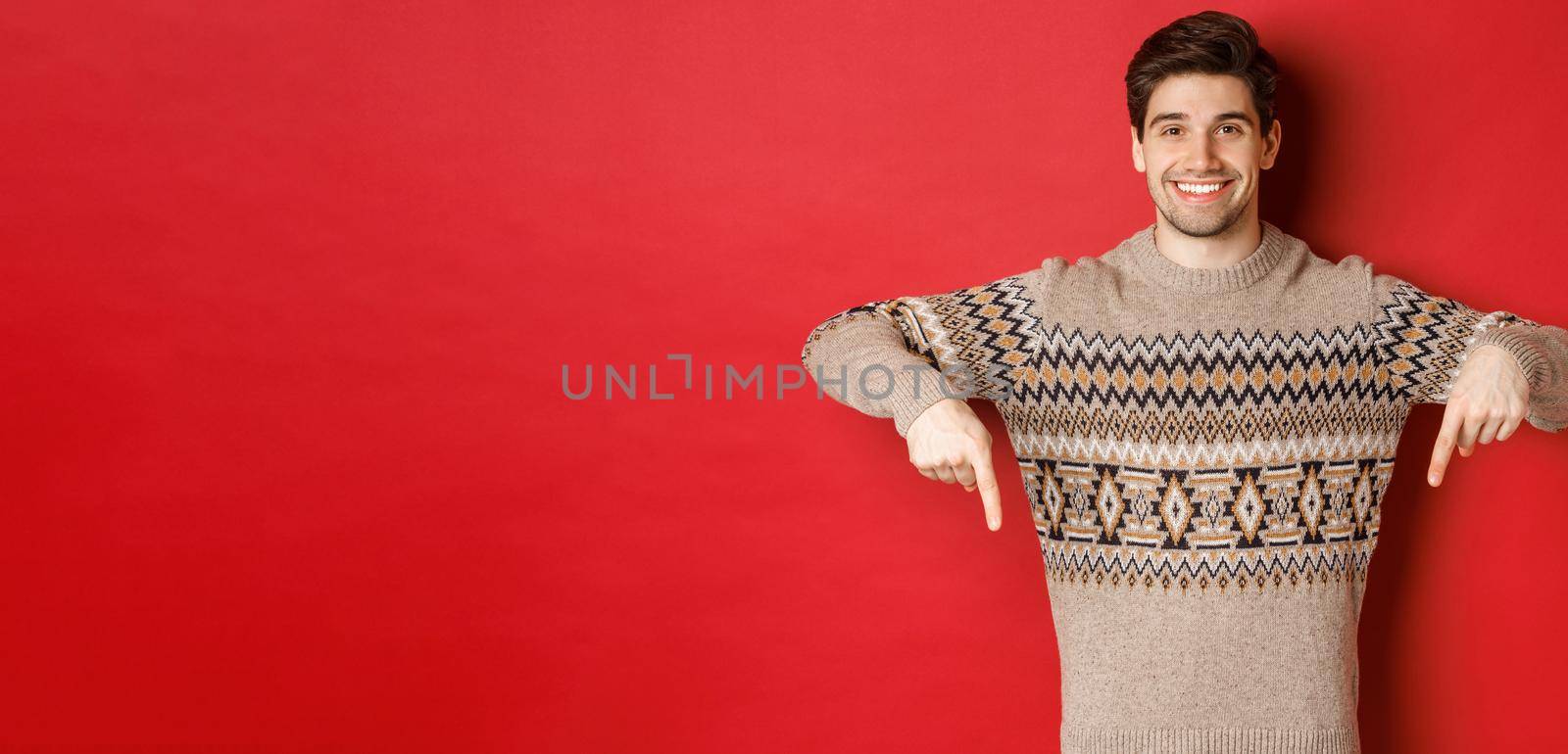 Concept of christmas celebration, winter holidays and lifestyle. Attractive happy man in xmas sweater showing promotion, pointing fingers down at logo, standing over red background.