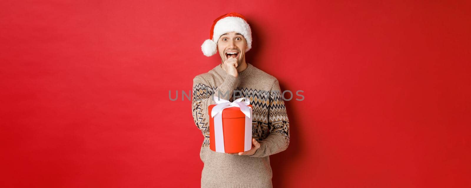 Image of handsome guy in santa hat and christmas sweater, excited to open xmas gift, looking amazed and holding present in one hand, standing over red background.