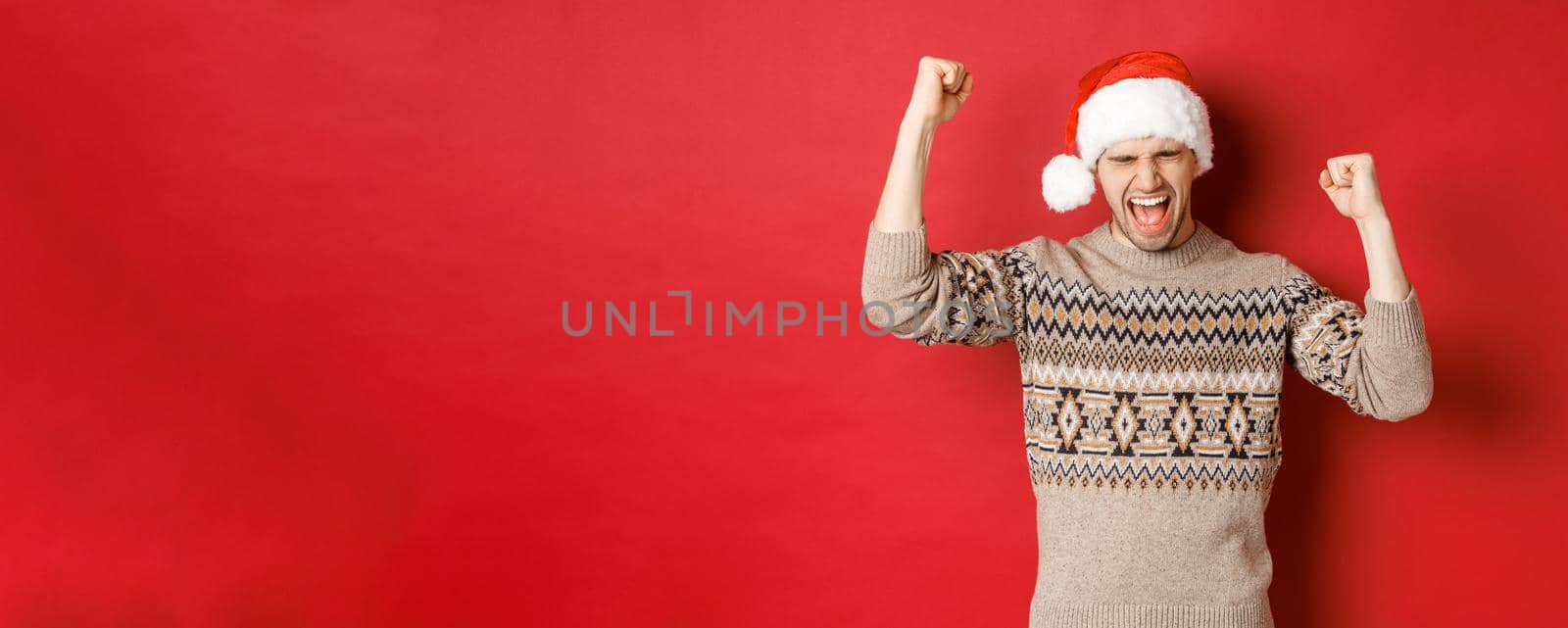 Satisfied handsome man, feeling lucky and happy, shouting for joy and making fist pumps, celebrating victory or win, receive awesome christmas gift, standing in santa hat over red background.