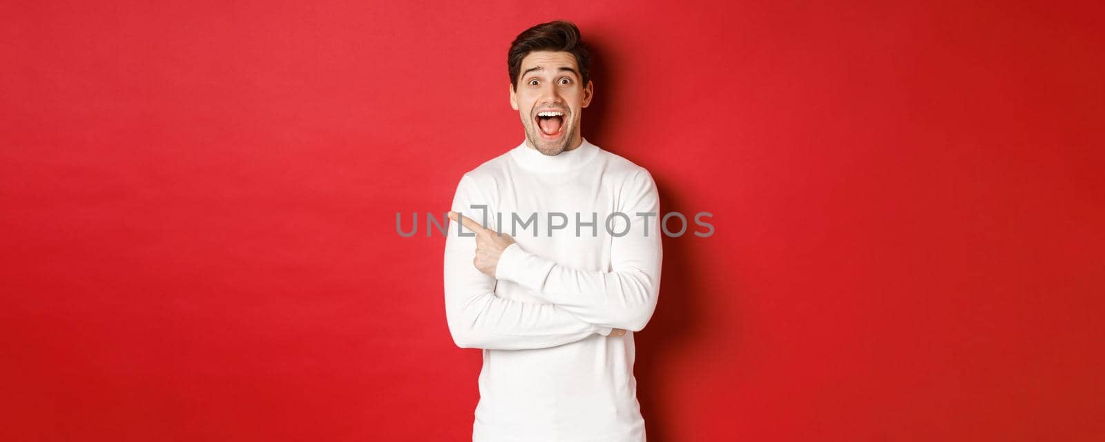 Concept of winter holidays, christmas and lifestyle. Amazed handsome man in white sweater showing logo, pointing finger left and looking excited, standing against red background by Benzoix