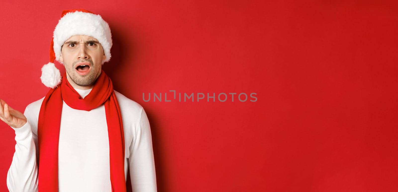 Concept of christmas, winter holidays and celebration. Portrait of confused man in santa hat and scarf, frowning and looking perplexed, standing over red background.