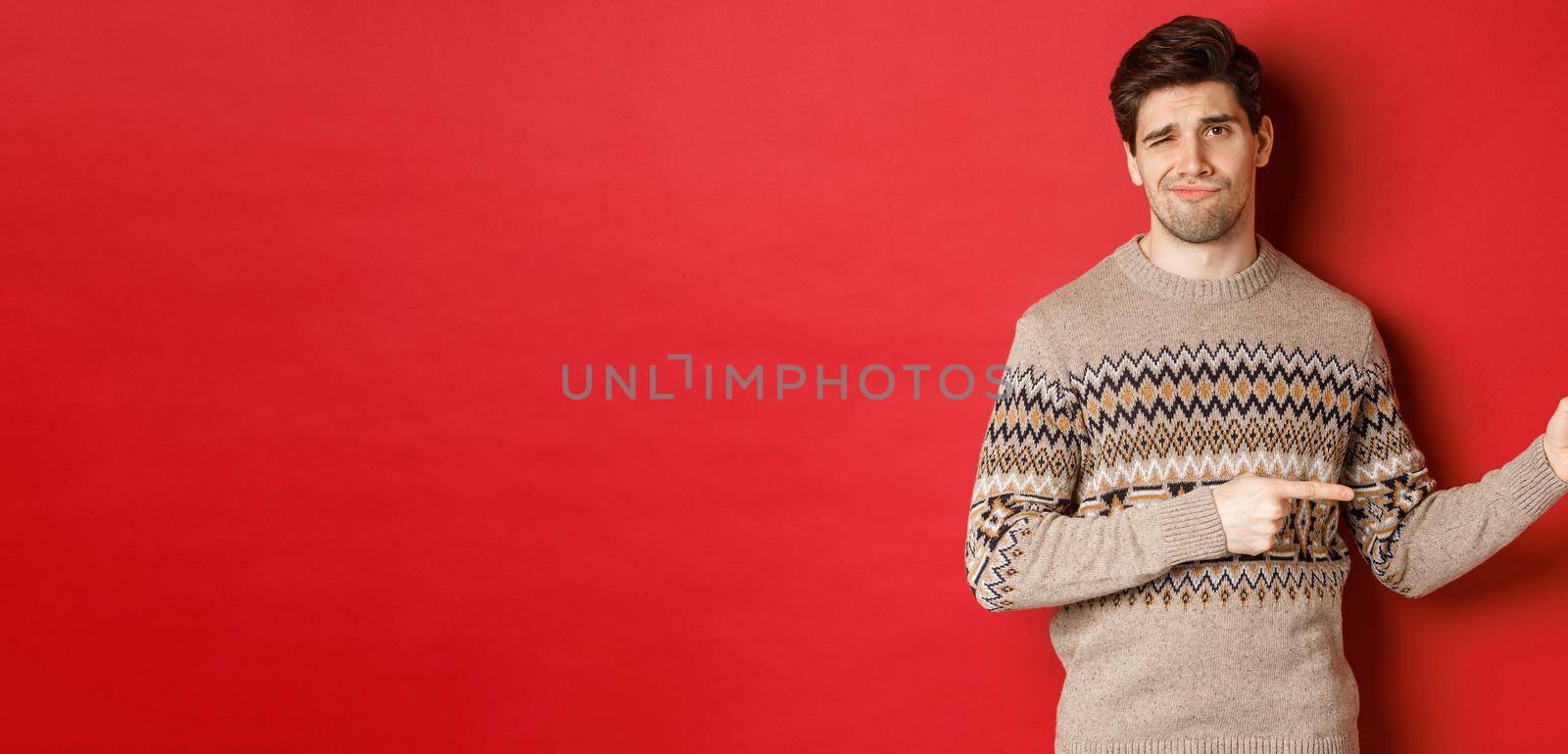 Concept of christmas celebration, winter holidays and lifestyle. Image of skeptical and displeased handsome man in xmas sweater, pointing fingers right and do not recommend product.