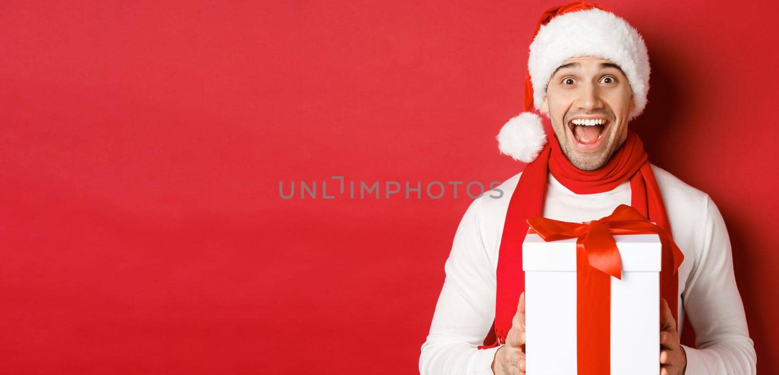 Concept of winter holidays, christmas and lifestyle. Close-up of excited handsome man in santa jat and scarf, looking amazed and receiving new year present, standing over red background.