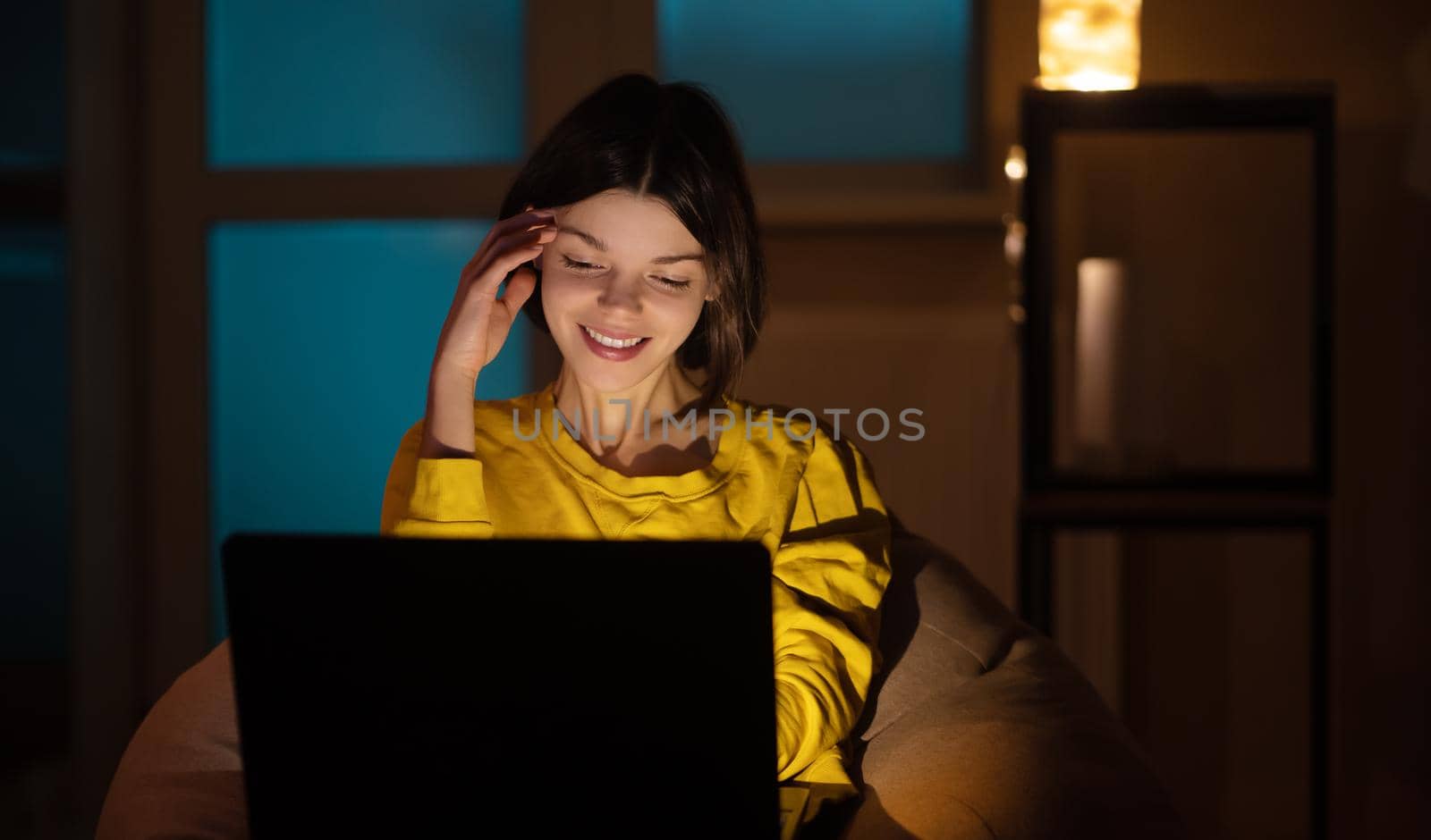 Portrait of beautiful caucasian girl uses laptop while sitting at home. In the evening creative woman works on a computer in her cozy living room.