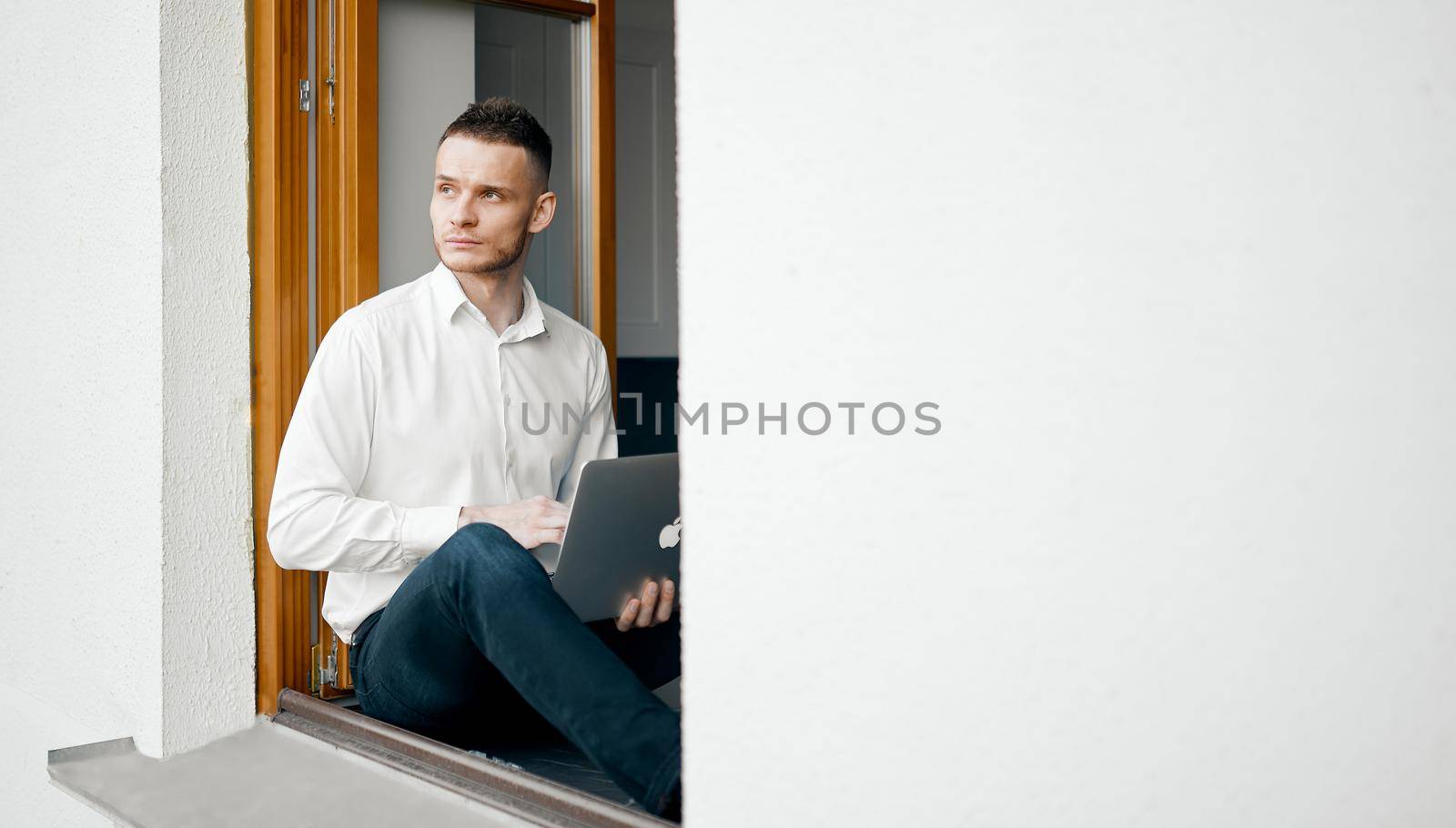 The guy is sitting on the windowsill of the house. Looks outside. A laptop in hand. High quality photo