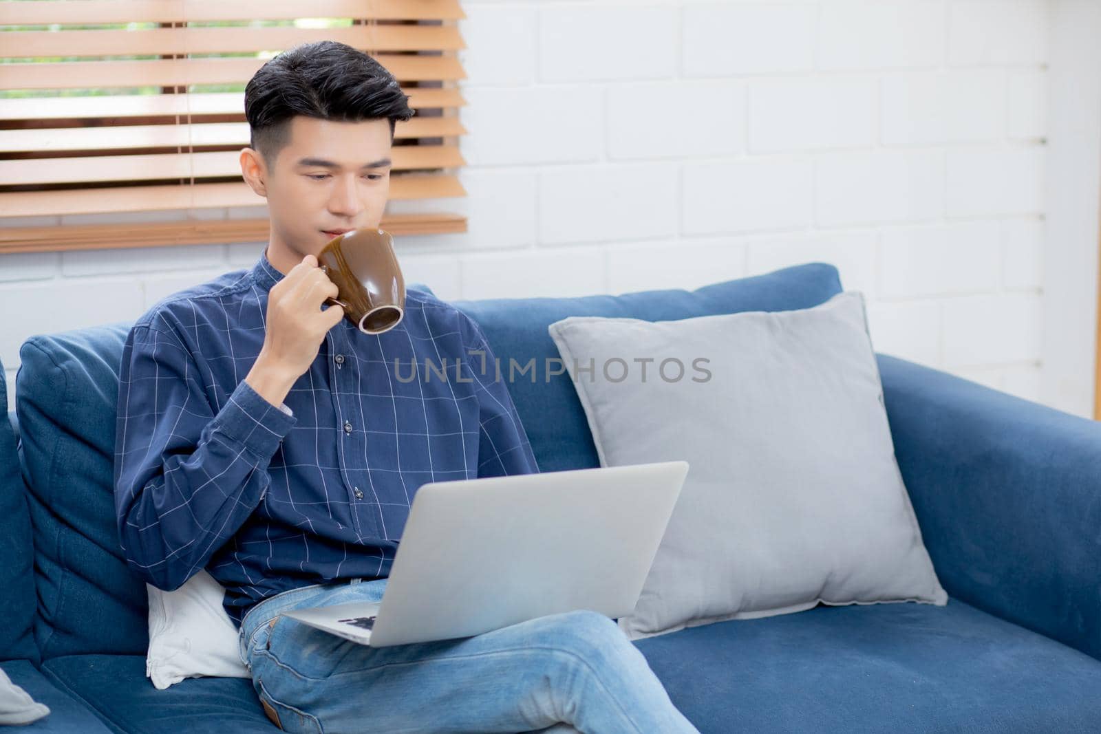Young asian businessman smile and work from home with laptop computer online on sofa in living room, freelance business man using notebook and drink coffee on couch, new normal, lifestyle concept. by nnudoo