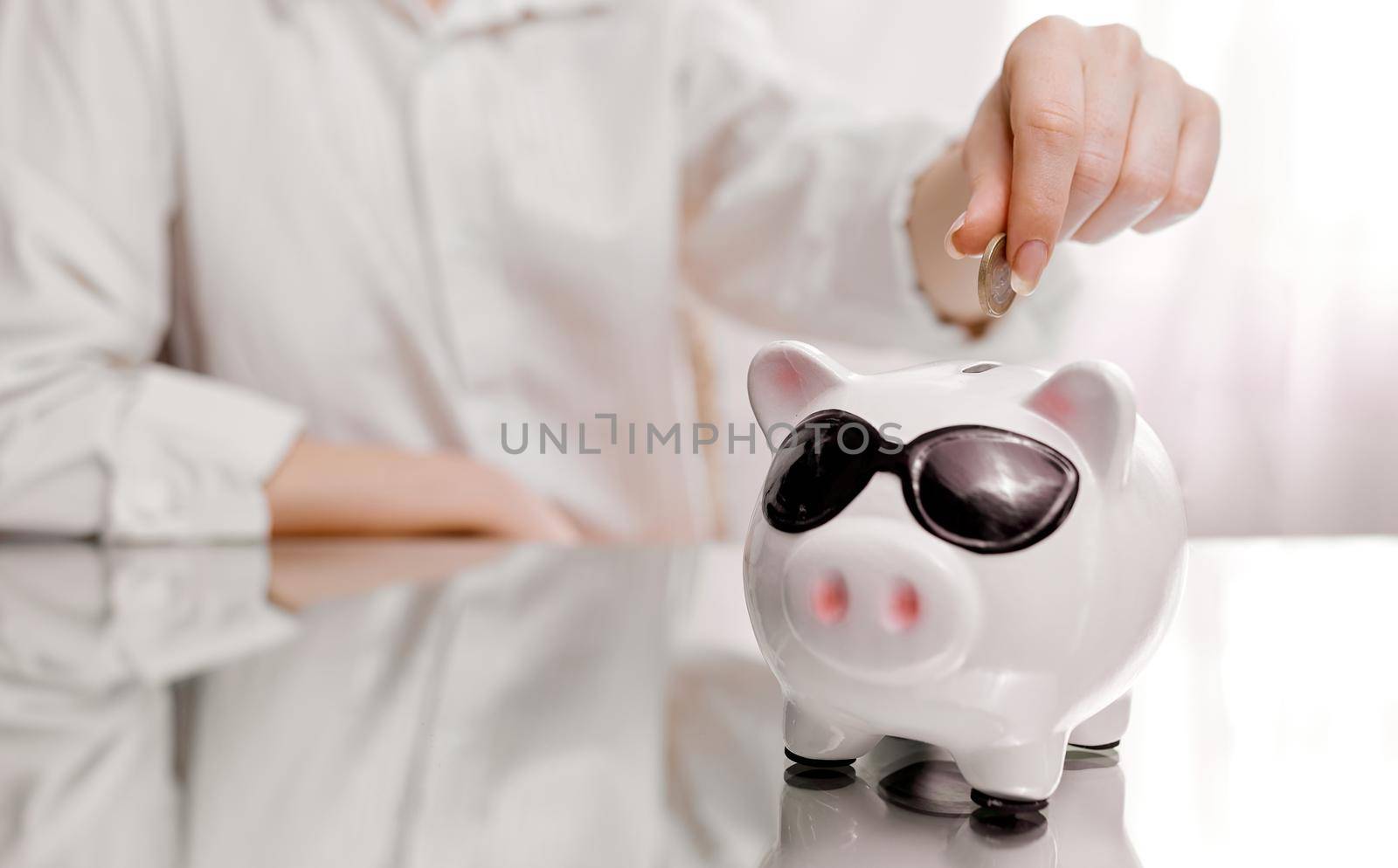 A woman puts a coin in a piggy bank on a mirrored table . High quality photo