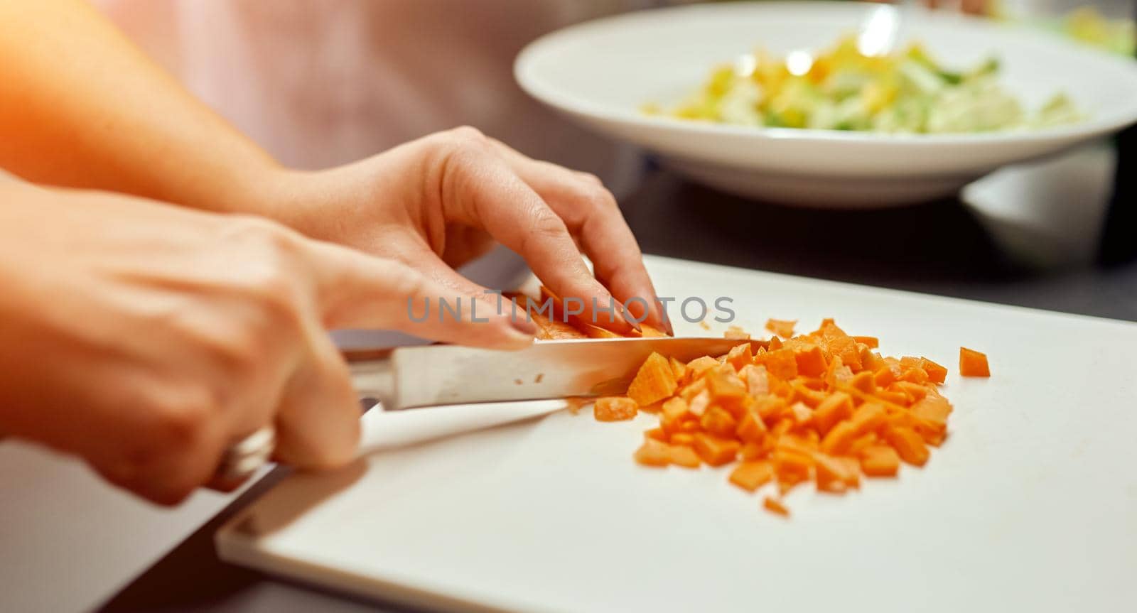 Woman cuts into small cubes on a plank carrots for salad, which stands against the background in a plate. High quality photo