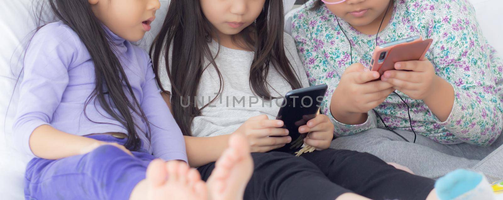 Happy asian little kid and friends playing smartphone on sofa at home, children using phone together on couch, girl watching smart phone for entertainment, lifestyle and communication concept. by nnudoo
