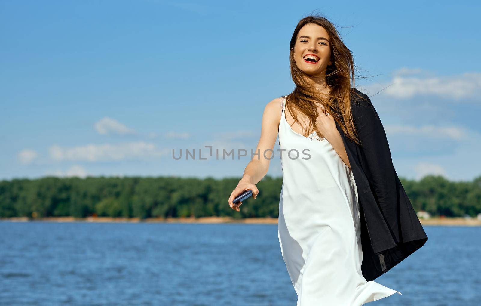Happy girl with phone in white dress with jacket by the sea. High quality photo