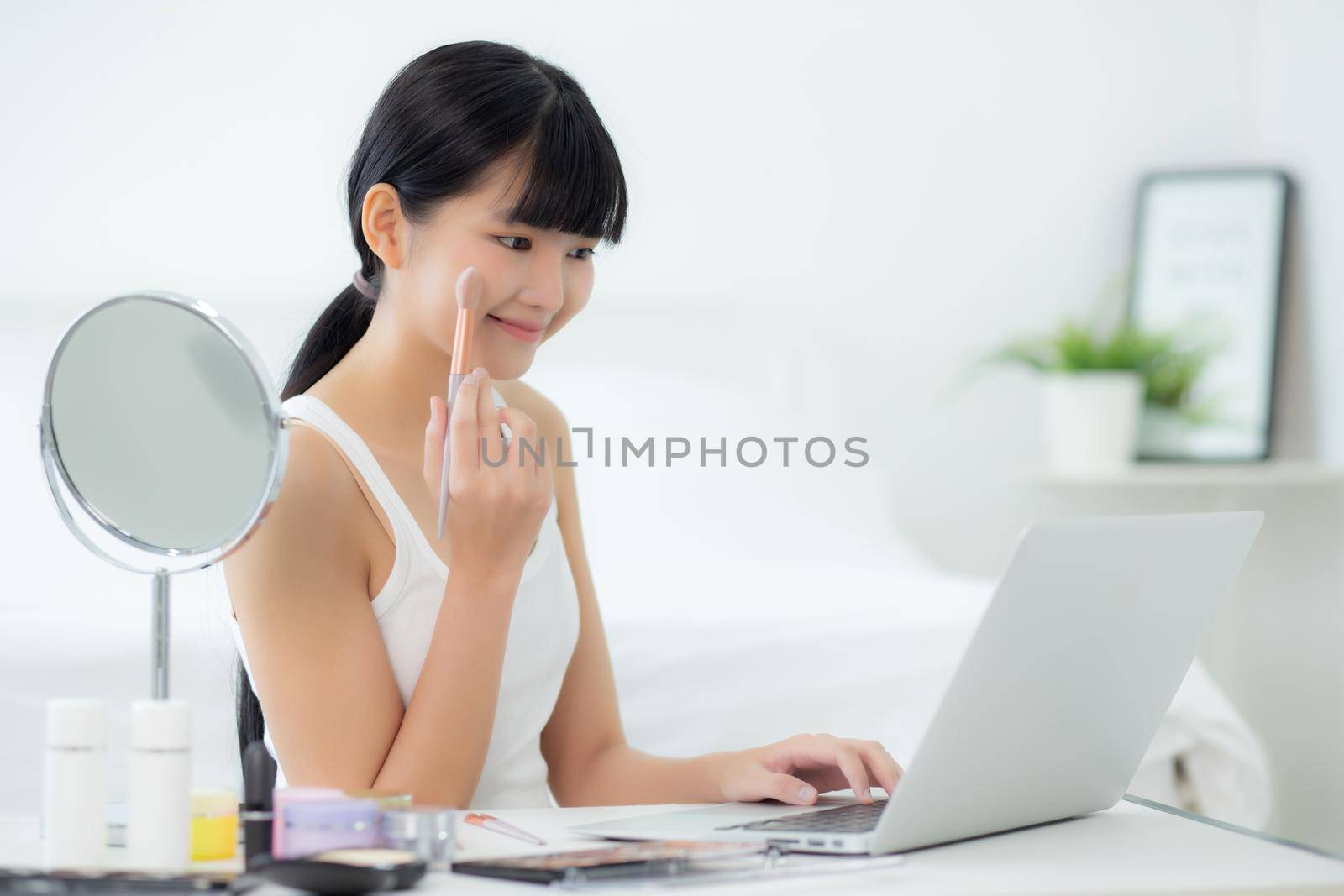 Beauty of young asian woman with learning makeup with brush on cheek on laptop computer with tutorial course online, female teaching and explain make up with cosmetic on streaming social media.