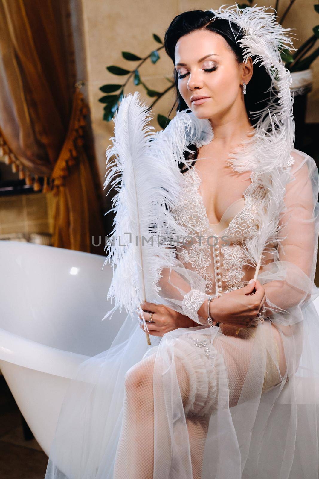 the bride, dressed in a boudoir transparent dress and underwear, sits on a vintage bathtub with a feather in her hands, The morning of the bride by Lobachad