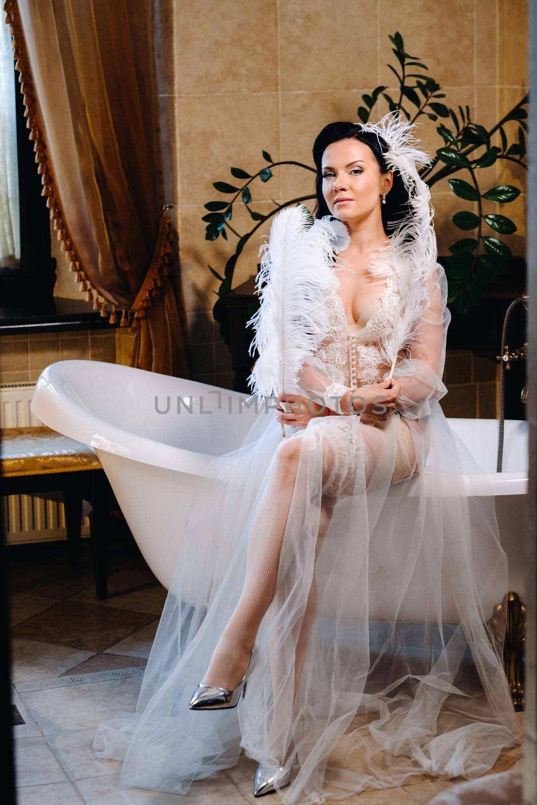 the bride, dressed in a boudoir transparent dress and underwear, sits on a vintage bathtub with a feather in her hands, The morning of the bride.