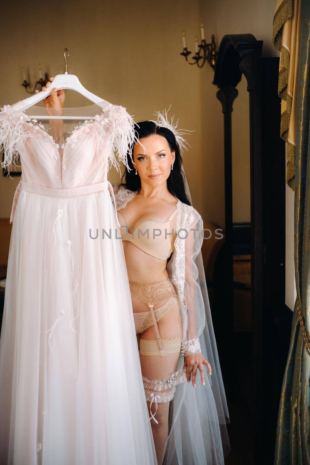 a bride dressed in a boudoir transparent dress and underwear holds her wedding dress in her hands in the interior of the house.
