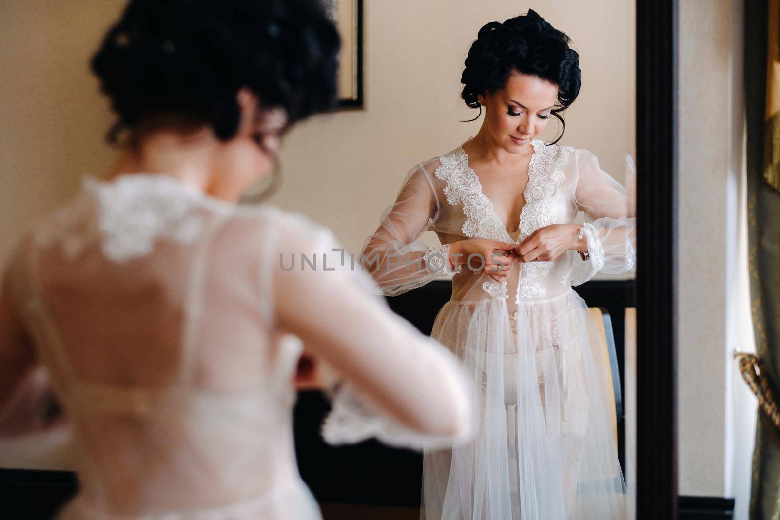 the bride, dressed in a boudoir transparent dress and underwear, stands at home in the morning by the mirror.