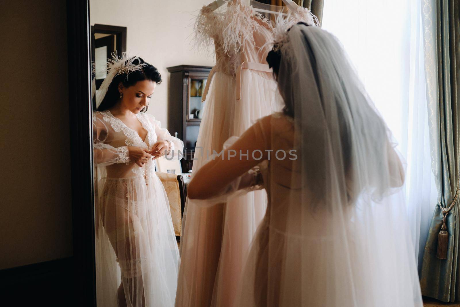 the bride, dressed in a boudoir transparent dress and underwear, stands at home in the morning by the mirror by Lobachad