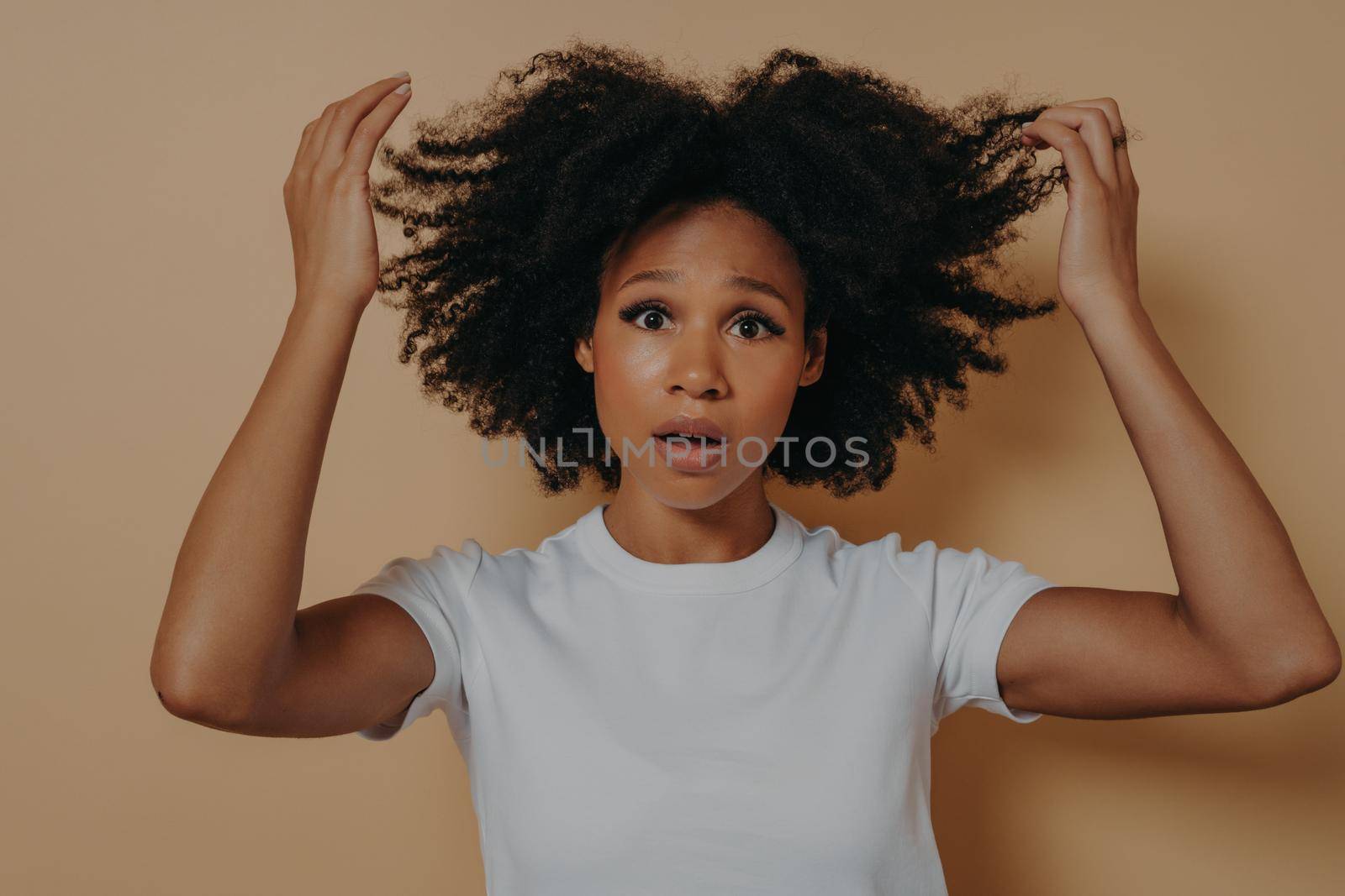 Worried young african american girl in white tshirt posing while standing isolated on beige studio background. Raising hands up in disbelief with shocked facial expression. Human emotions concept