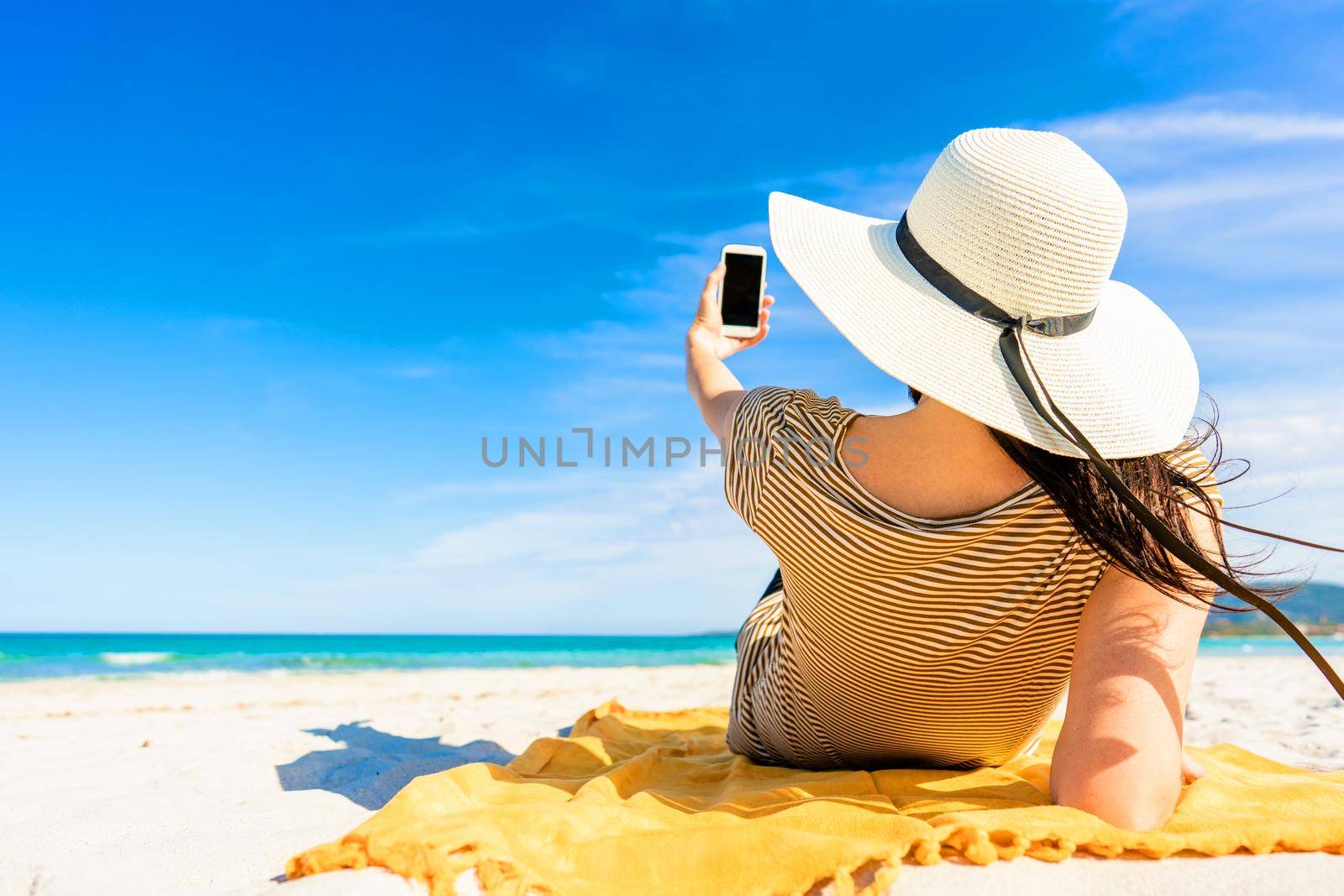 Back shot of fashionable young unrecognizable woman in large white hat lying on sand of tropical beach taking selfie with smartphone sharing vacations on social network. Bright vivid color photograph