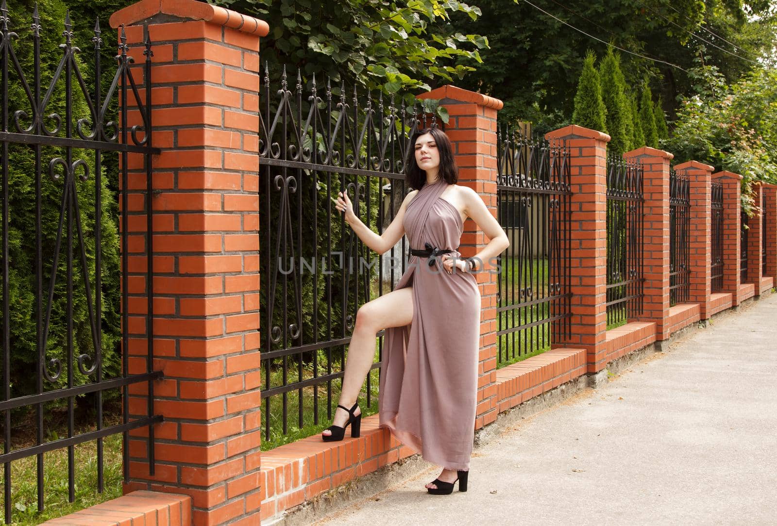 young beautiful brunette woman in beige dress standing on the sidewalk near metal fence on summer day