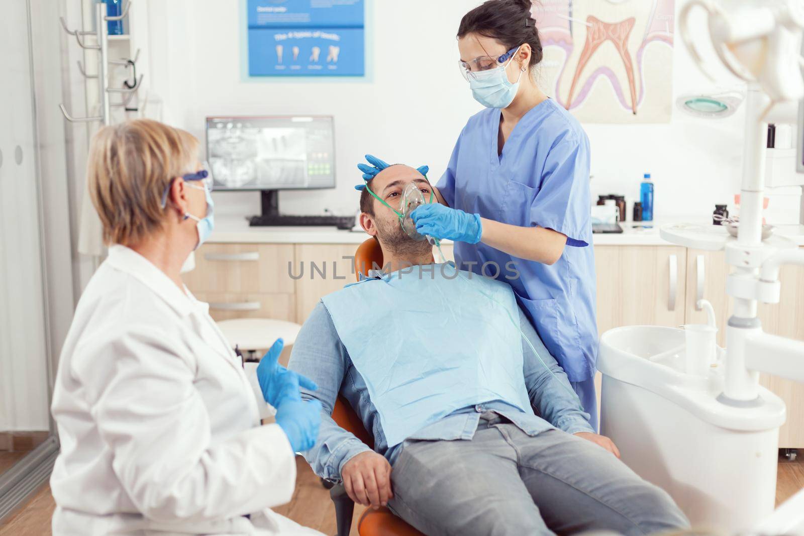 Stomatologist nurse putting oxigen mask before tooth surgery sitting on dental chair in stomatology cabinet. Senior doctor and nurse working in modern orthodontic office wearing mask and gloves
