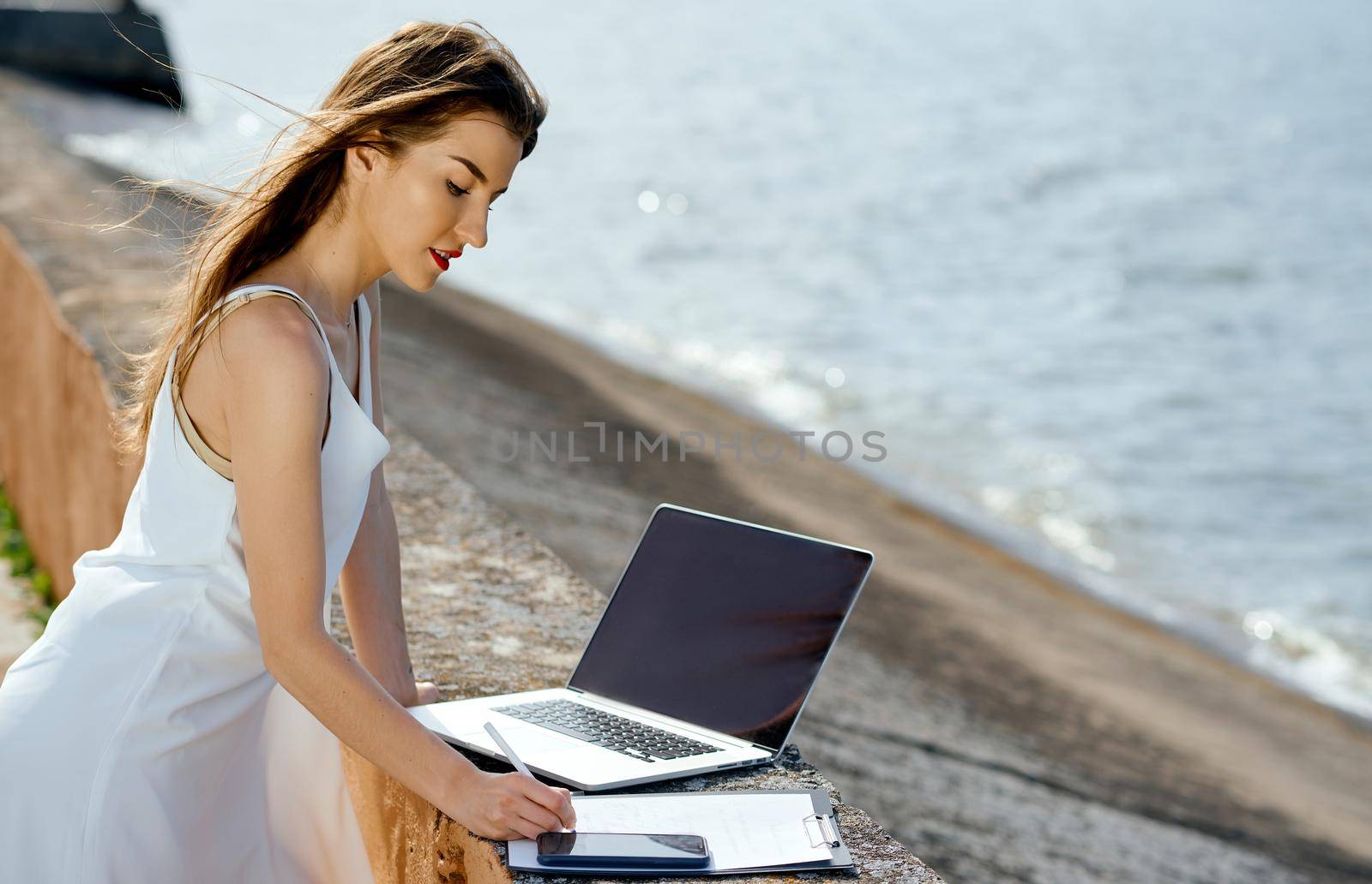 beautiful, serious woman in a white dress with a tablet, pen, laptop phone on the dock by AntonIlchanka