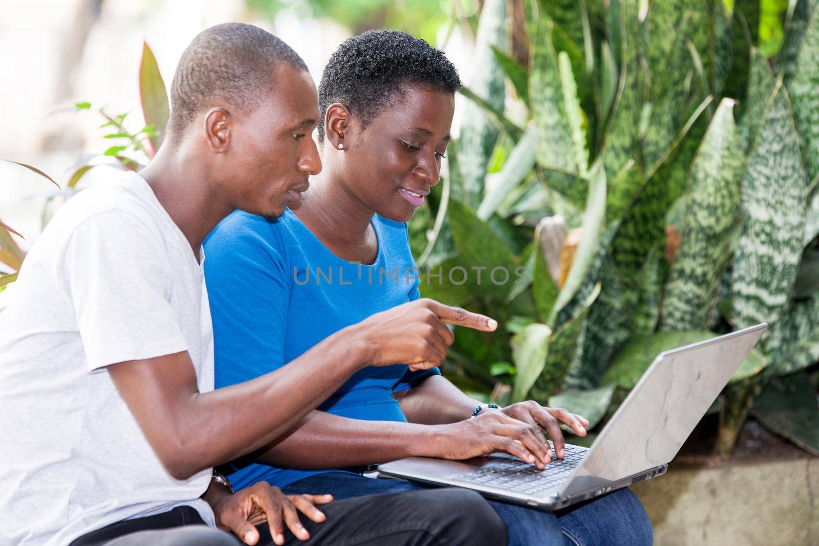 young students sitting in park with laptop doing research while smiling.