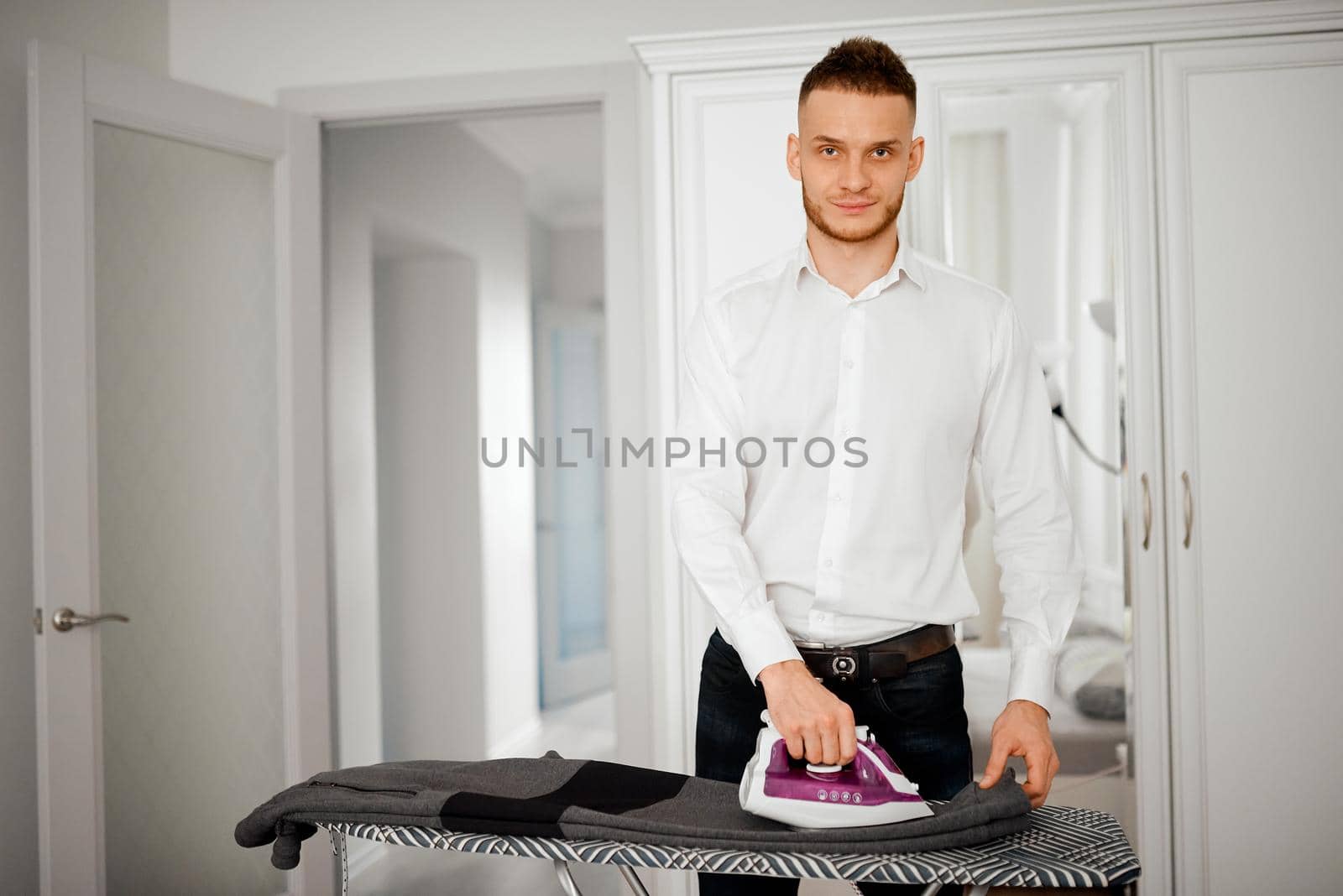 A man irons a jacket with an iron in the apartment and looks into the camera. High quality photo