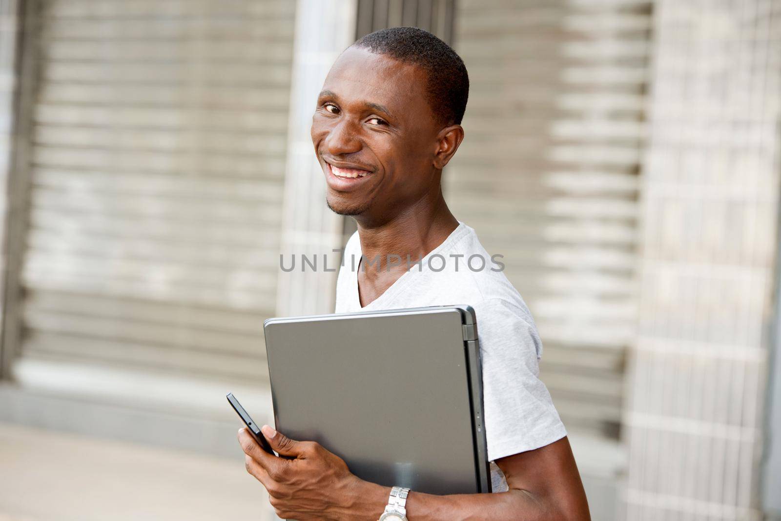 Young student standing in front of study room with laptop and looking at the camera smiling.