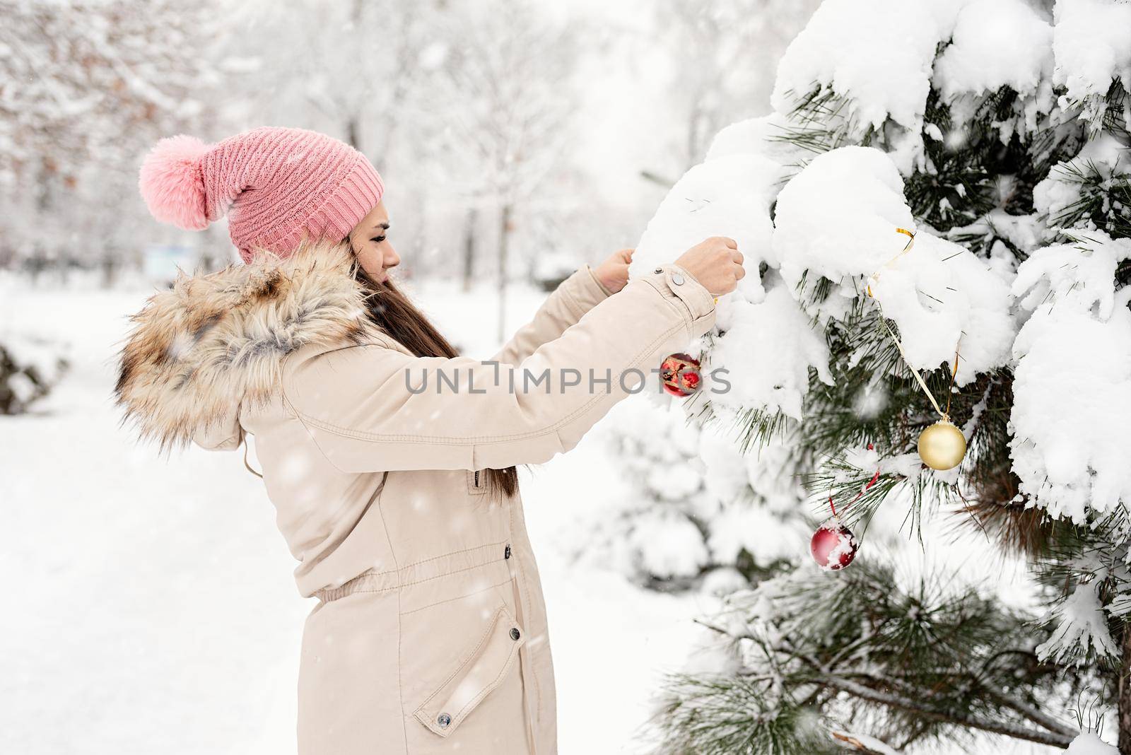 Merry Christmas and happy New Year. Beautiful woman in warm winter clothes decorating Christmas tree in a park in snowy day