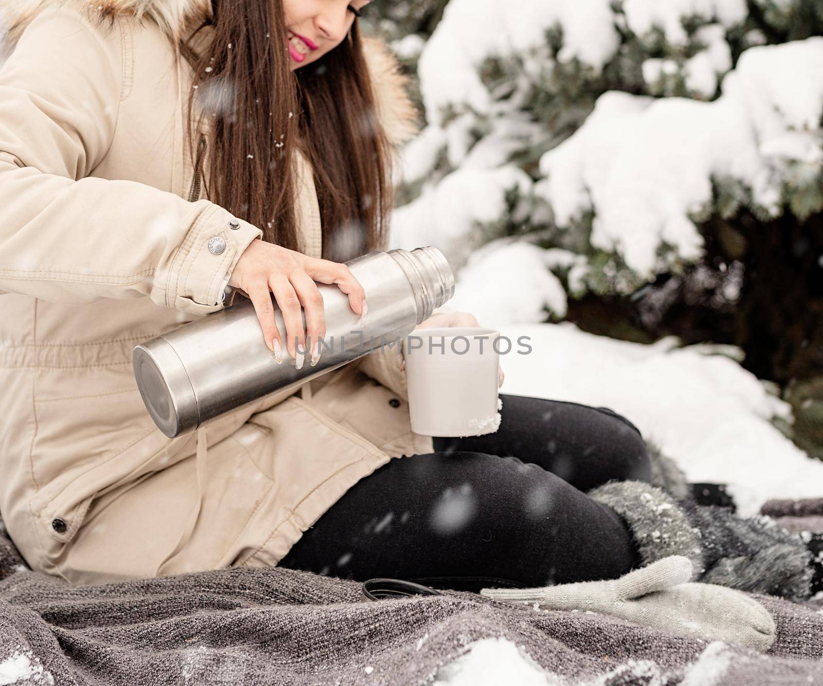 Beautiful woman in warm winter clothes holding thermos and drinking hot tea or coffee outdoors in snowy day by Desperada