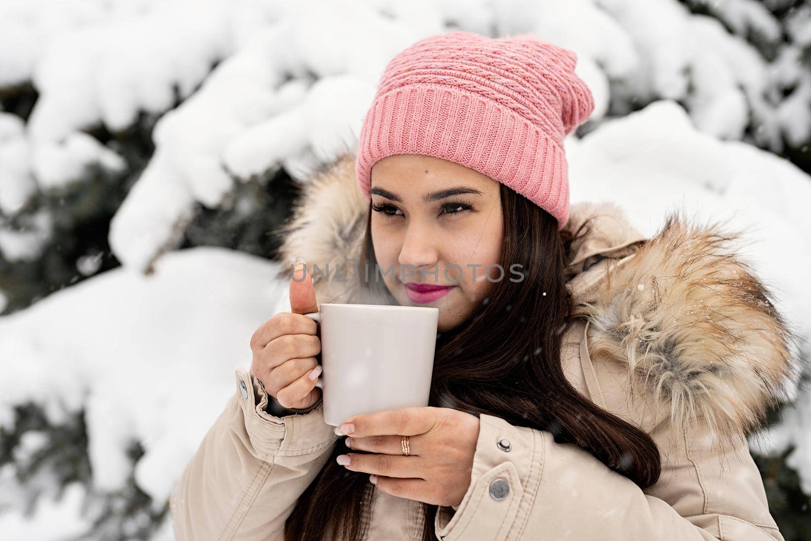 Beautiful woman in warm winter clothes holding cup drinking hot tea or coffee outdoors in snowy day by Desperada