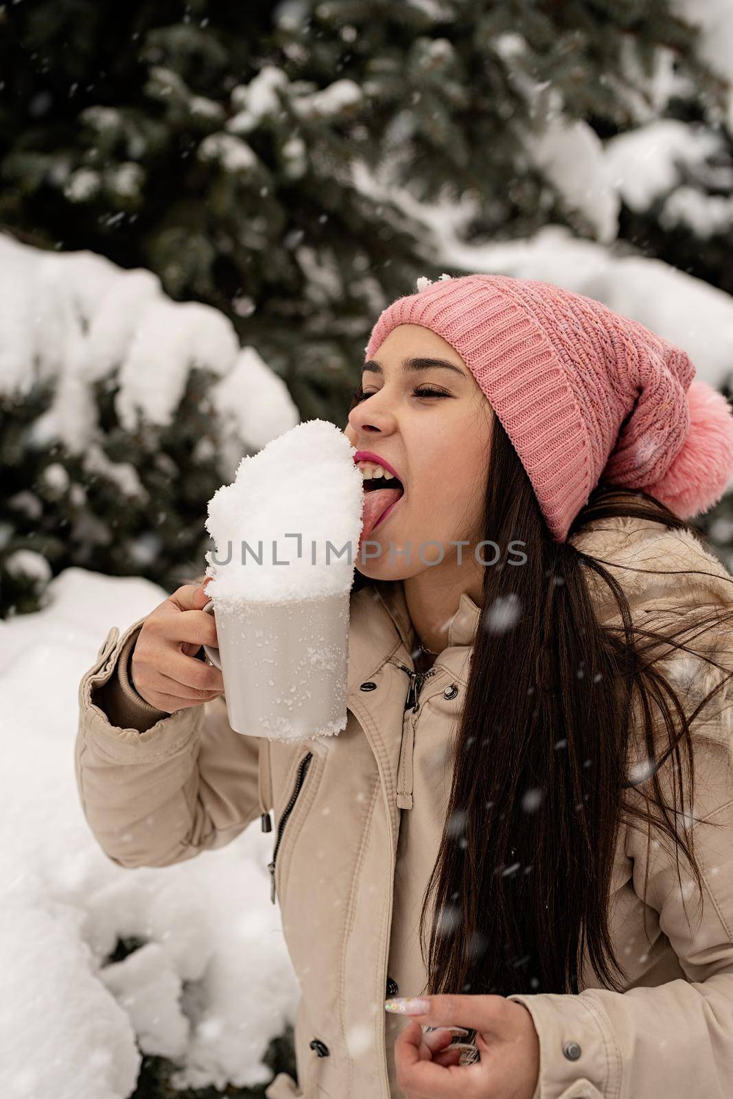 woman in warm winter clothes standing by the big christmas tree outdoors and licking snow from the cup by Desperada