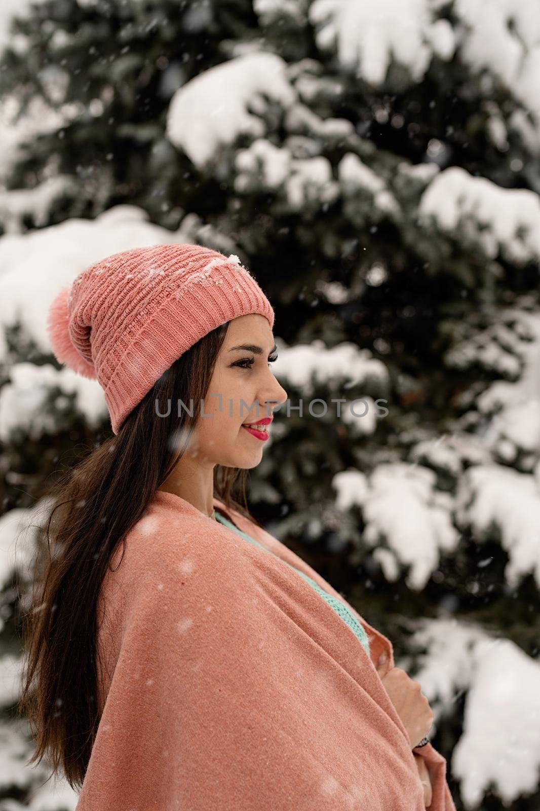 Merry Christmas and happy New Year. Portrait of beautiful brunette woman in pink warm winter clothes standing outdoors in snowy day by christmas tree , snow falling