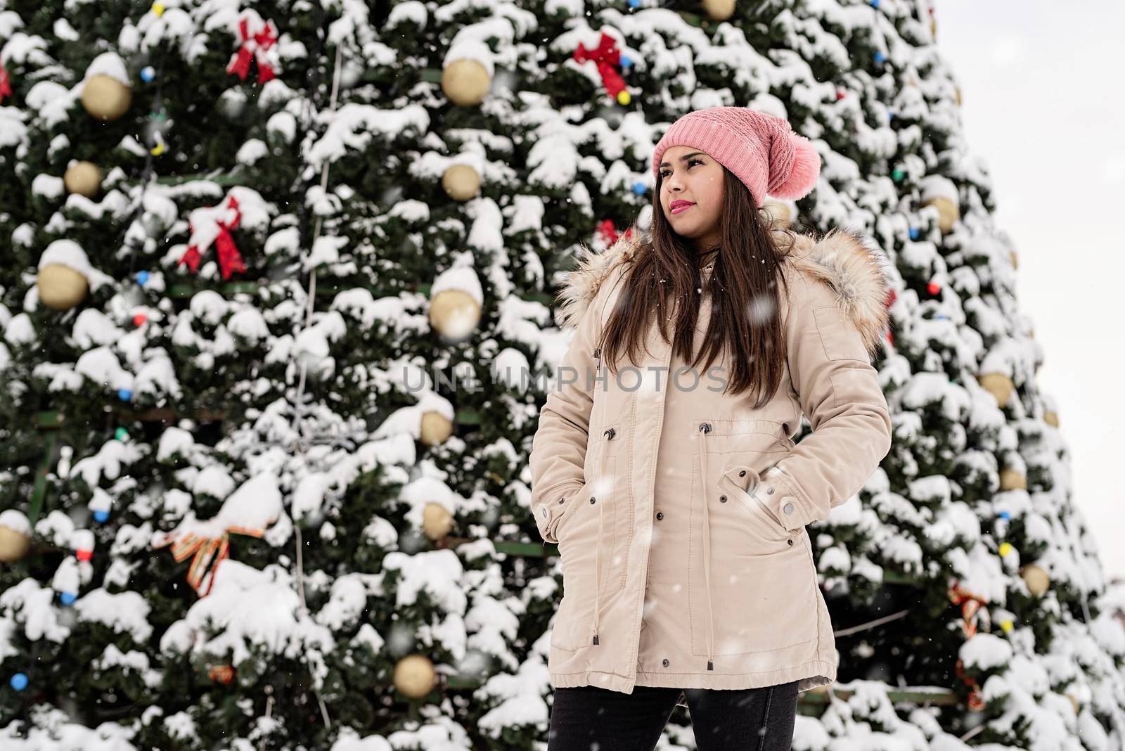 Woman in warm winter clothes standing by the big christmas tree outdoors, snow falling by Desperada