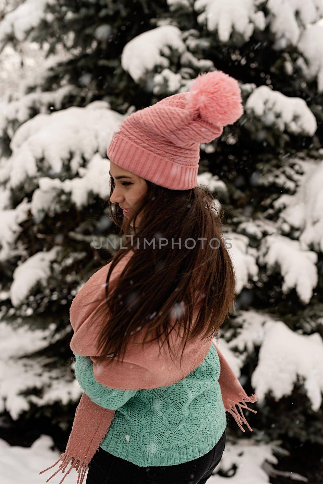 Merry Christmas and happy New Year. Portrait of beautiful brunette woman in pink warm winter clothes standing outdoors in snowy day by christmas tree shaking hair, snow falling