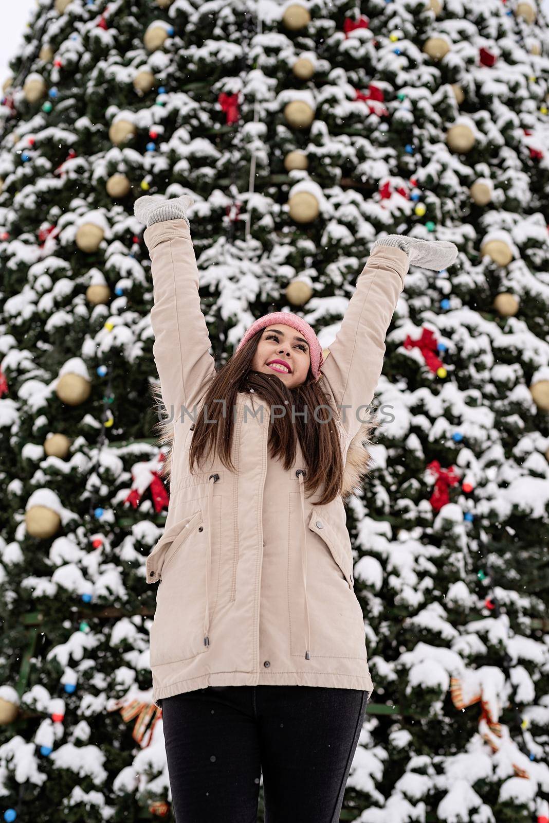 Merry Christmas and happy New Year. Happy Woman in warm winter clothes standing by the big christmas tree outdoors, arms up, snow falling