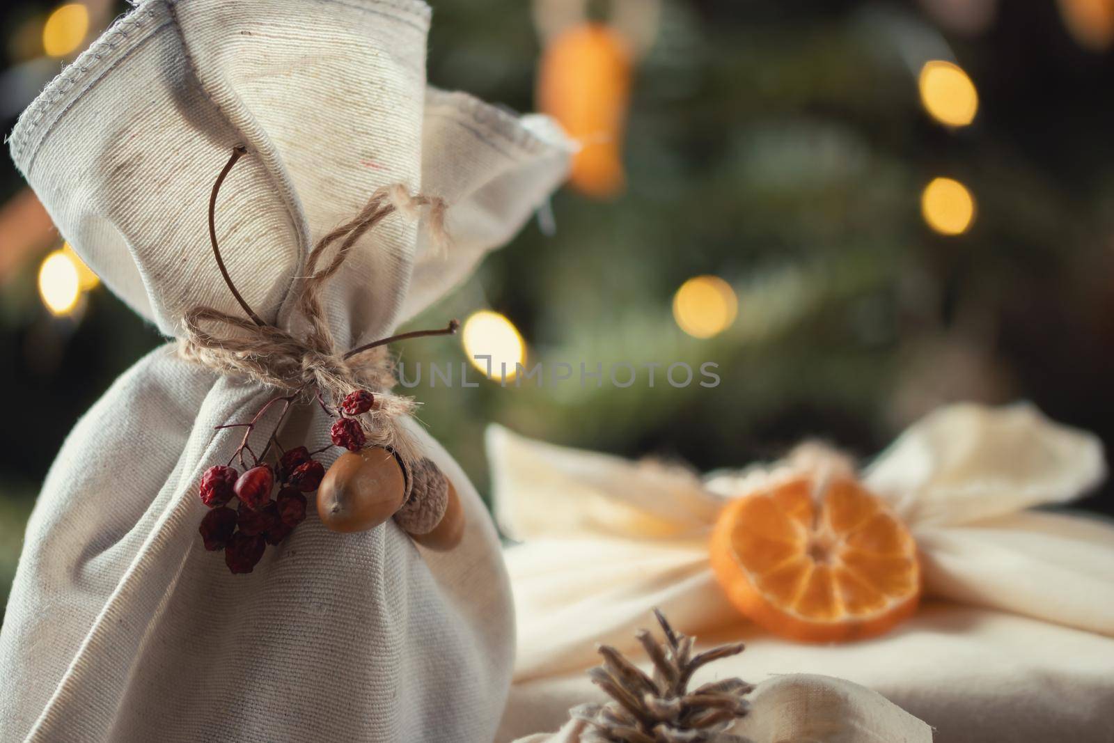 Zero waste christmas concept. Packed in natural fabric gifts and decorations from natural materials on wooden table near Christmas tree with lights.