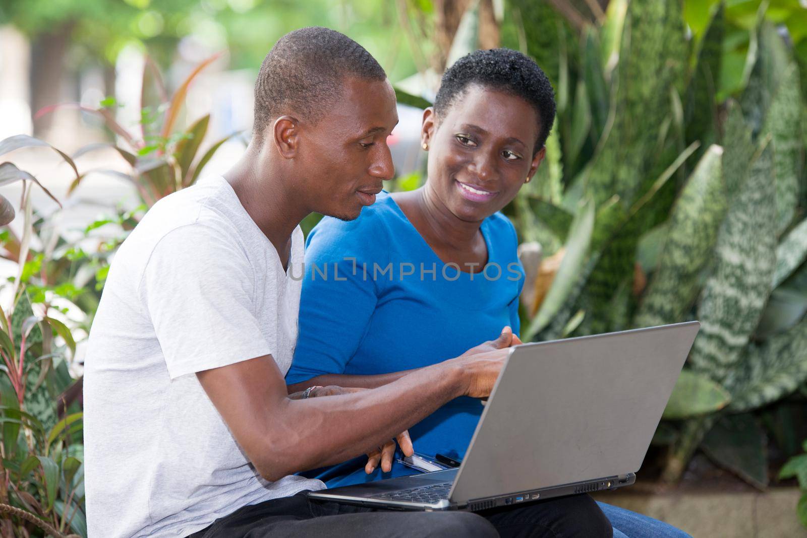 young students sitting in park doing research looking at laptop while smiling.