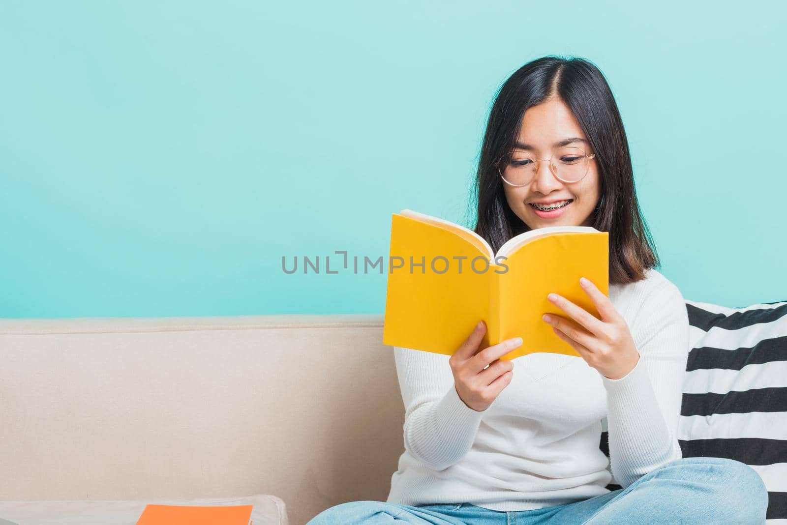 Asian beautiful young woman wearing eyeglasses sitting on sofa reading storybook, portrait relaxation of happy female smiling in living read a book studio shot isolated on blue background