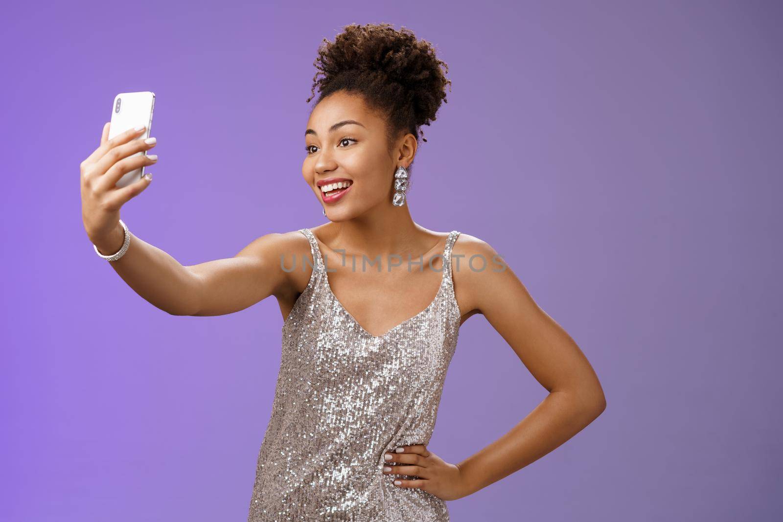 Confident glad attractive stylish millennial african-american woman in silver glittering dress taking selfie posing cute sassy hold hand waist extend arm record video message during night-out by Benzoix