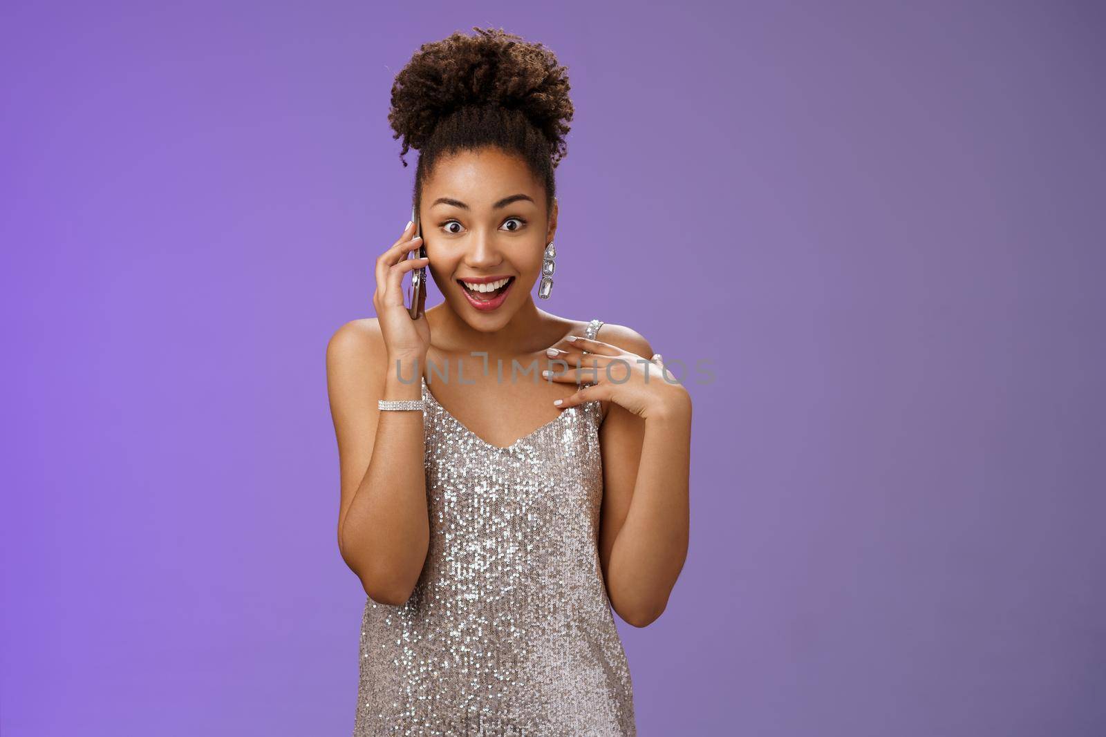 Impressed talkative charming african-american woman in shiny silver evening dress pointing herself amused surprised smiling talking smartphone widen eyes astonished, standing blue background.