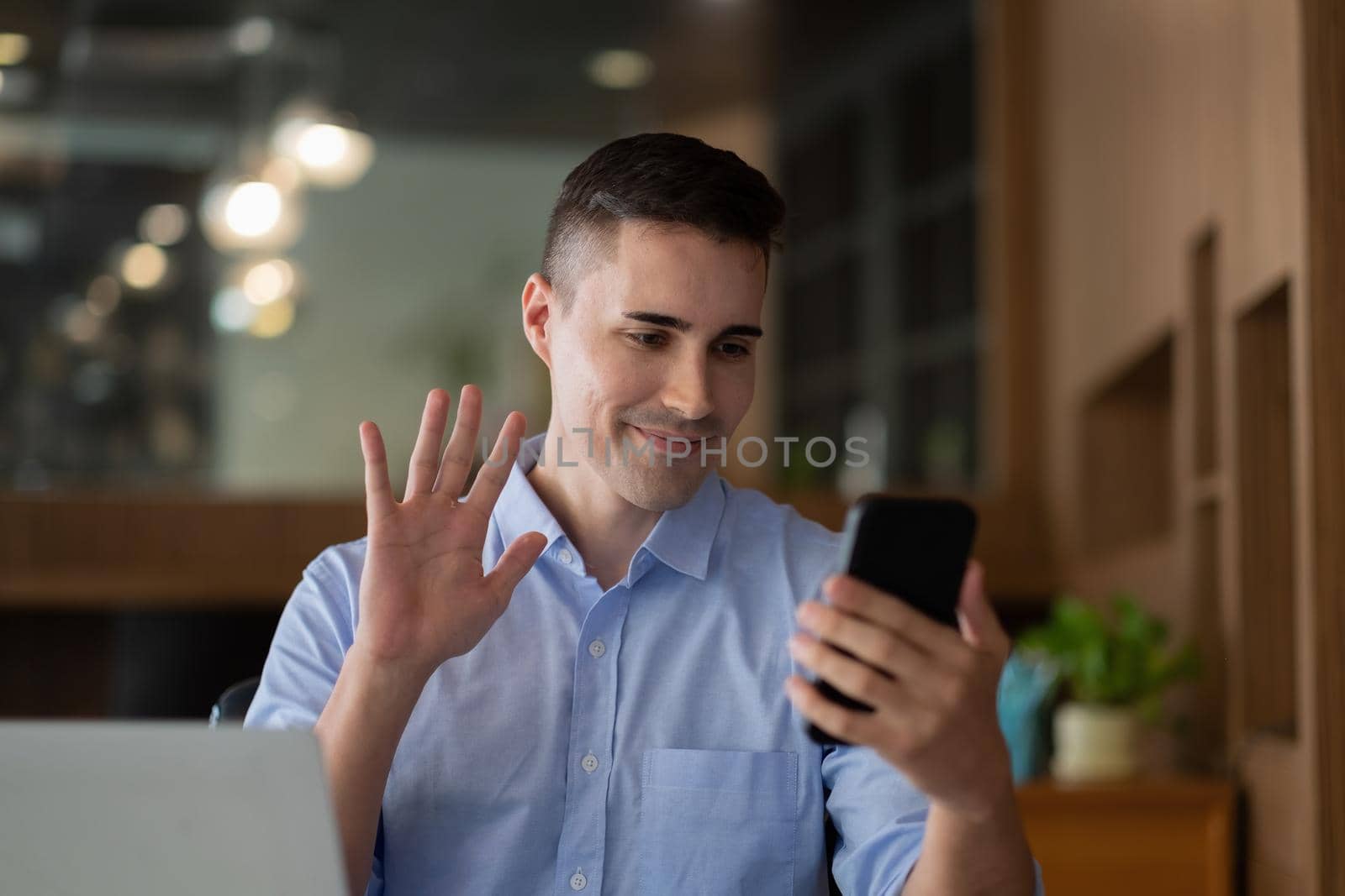 Cheerful happy businessman using smartphone app for virtual communication, holding phone in hand and waving at webcam, holding video call sitting at the desk. by nateemee