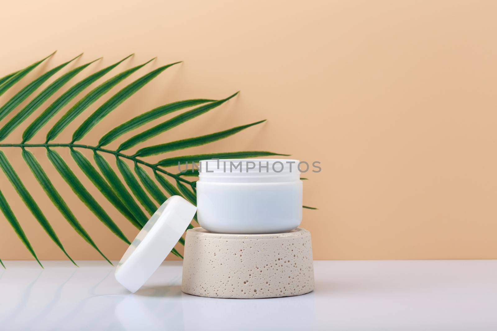 White opened cosmetic jar on gypsum podium against pastel beige background with palm leaf and copy space by Senorina_Irina