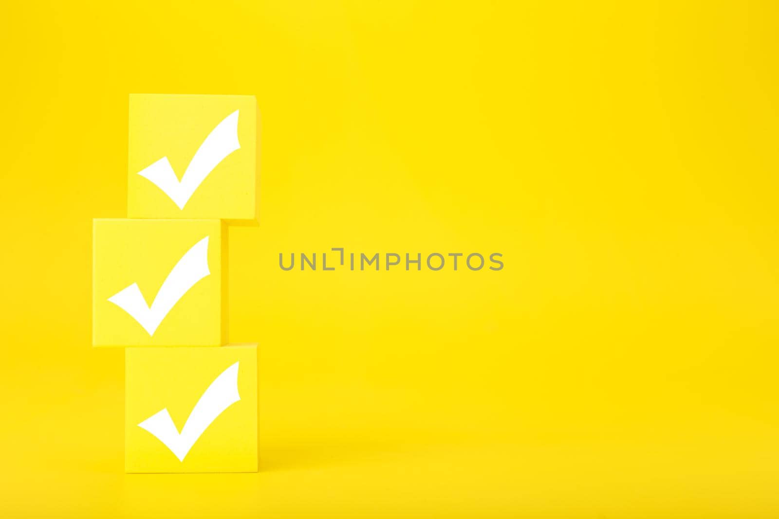 Three white checkmarks on stack of yellow toy cubes on bright yellow background with copy space. Concept of questionary, kids related checklist, to do list, planning, business or verification. 