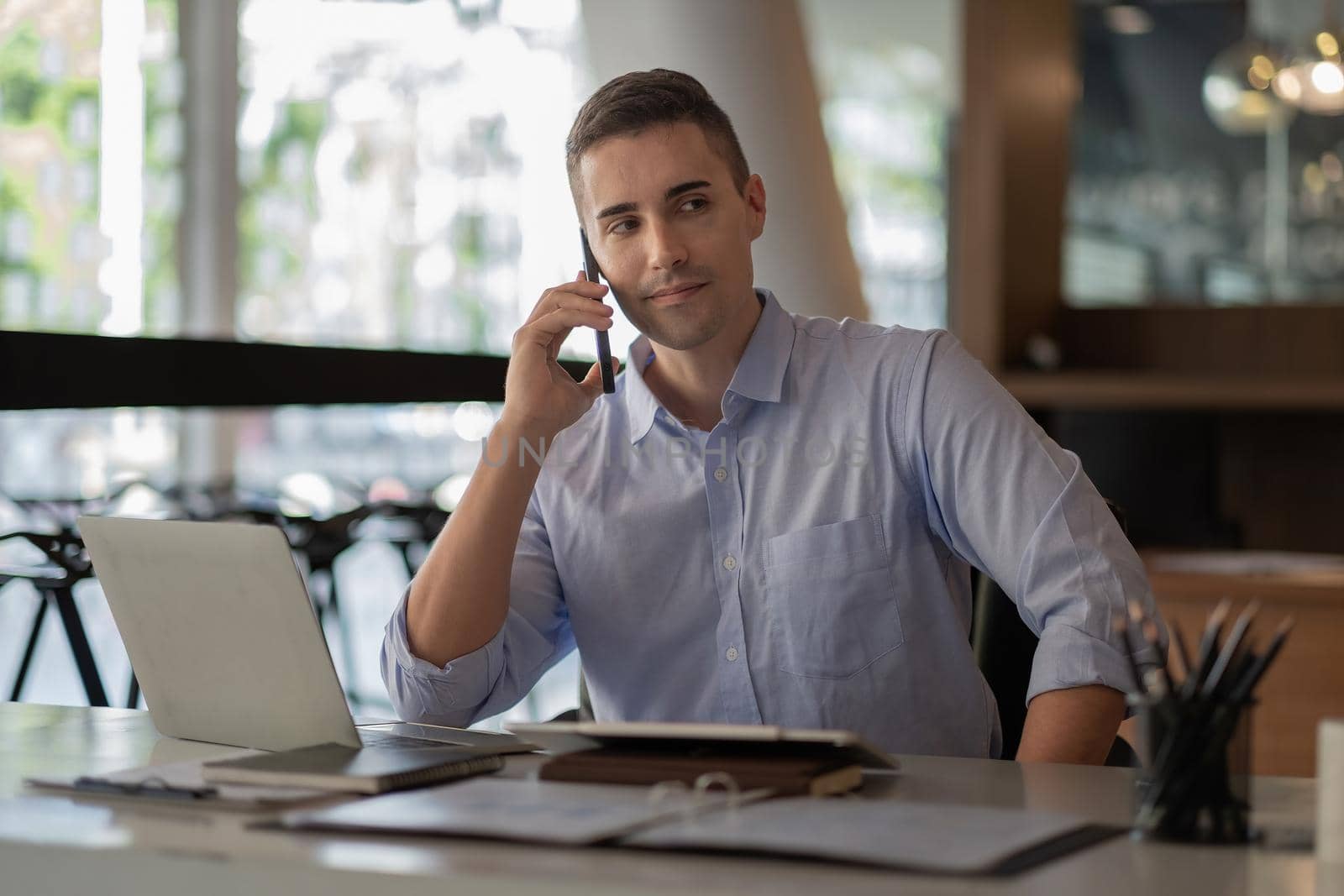 Portrait of young smiling cheerful entrepreneur in modern office making phone call while working with laptop