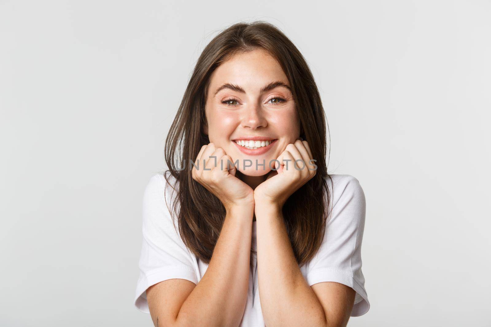 Close-up of silly and cute smiling brunette girl leaning on hands and looking with admiration, white background.