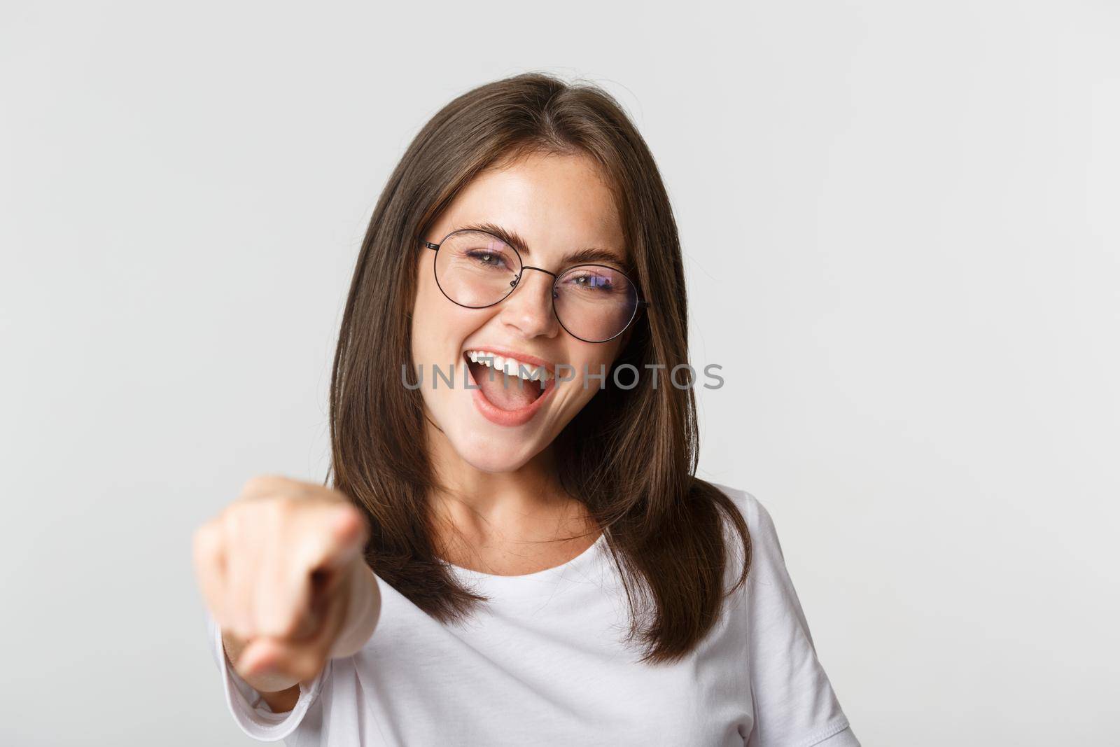 Close-up of cheerful beautiful girl in glasses smiling, poiting finger at camera.