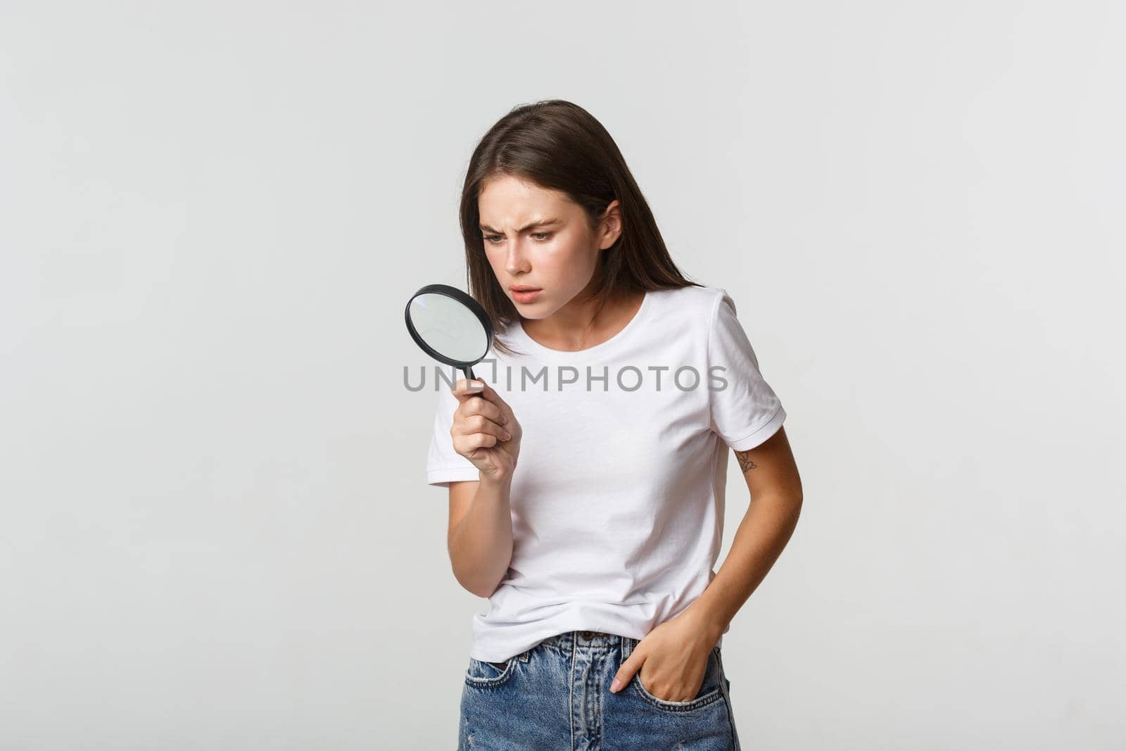 Serious-looking smart beautiful girl looking through magnifying glass down, searching for something.
