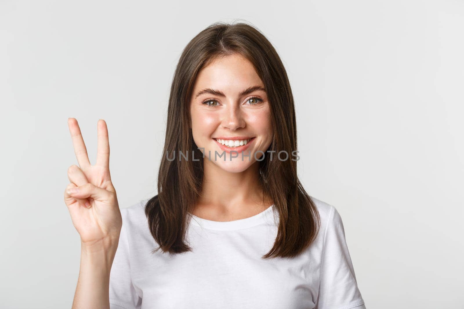 Portrait of beautiful and happy brunette girl smiling and showing peace gesture, standing white background.