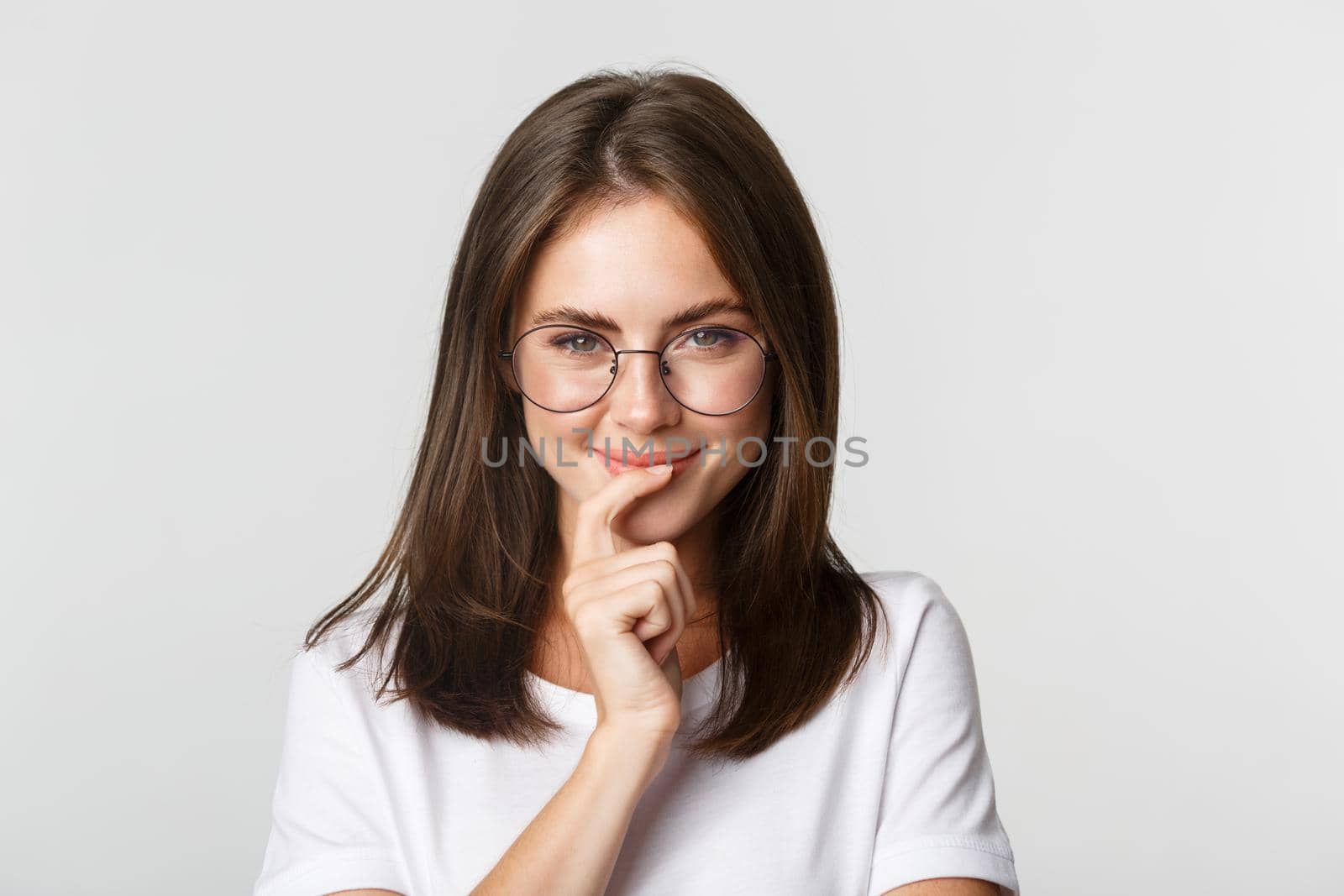 Close-up of thoughtful young smart girl in glasses smiling and looking intrigued, choosing product, thinking over white background.